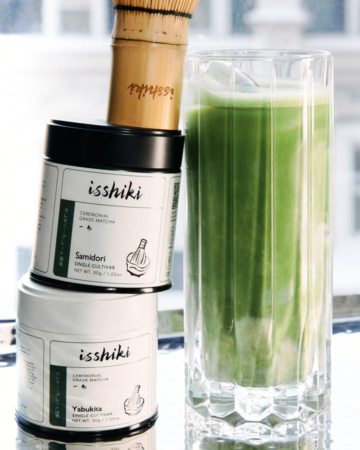 Ceremonial matcha powder jars with black lids next to a glass filled with green matcha.