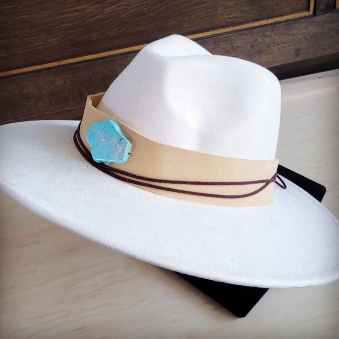 White wide-brim suede hat with tan hatband, turquoise concho, and dark cords