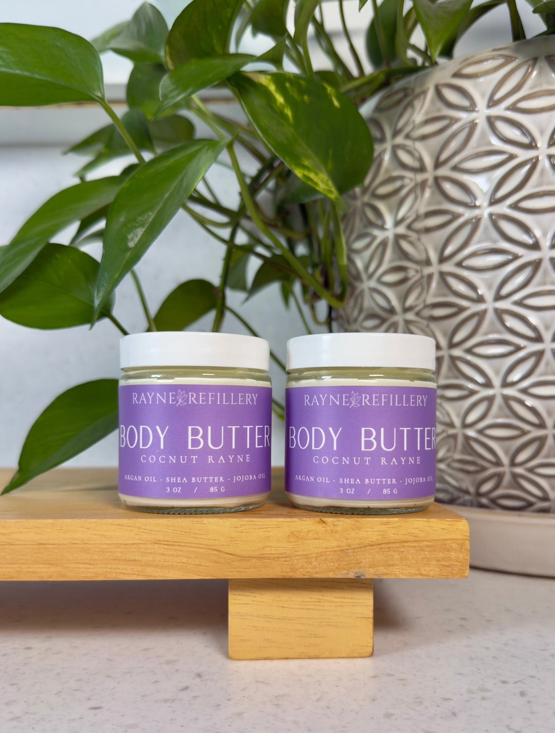 Two body butter jars with purple labels on a wooden stand beside green leaves