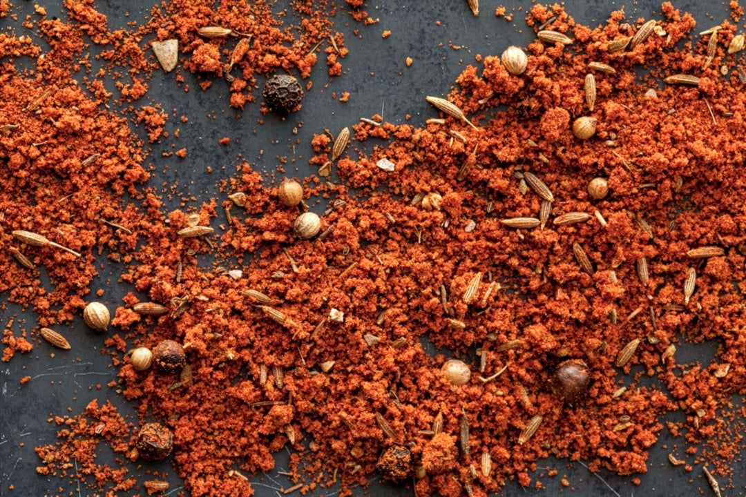 Ground red BBQ rub spice blend with herbs and seeds scattered on a dark surface