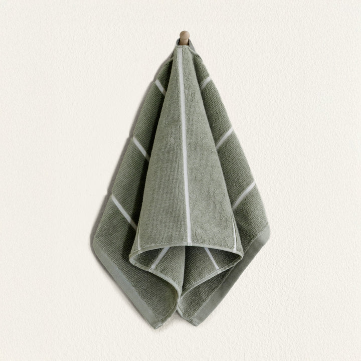 Sage green cotton towel with lighter border, folded and hanging on a small loop.