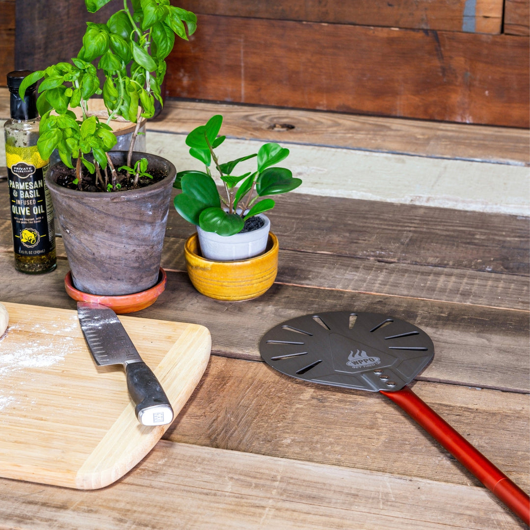 Round pizza peel with red handle and turning disc on a wooden table with herbs