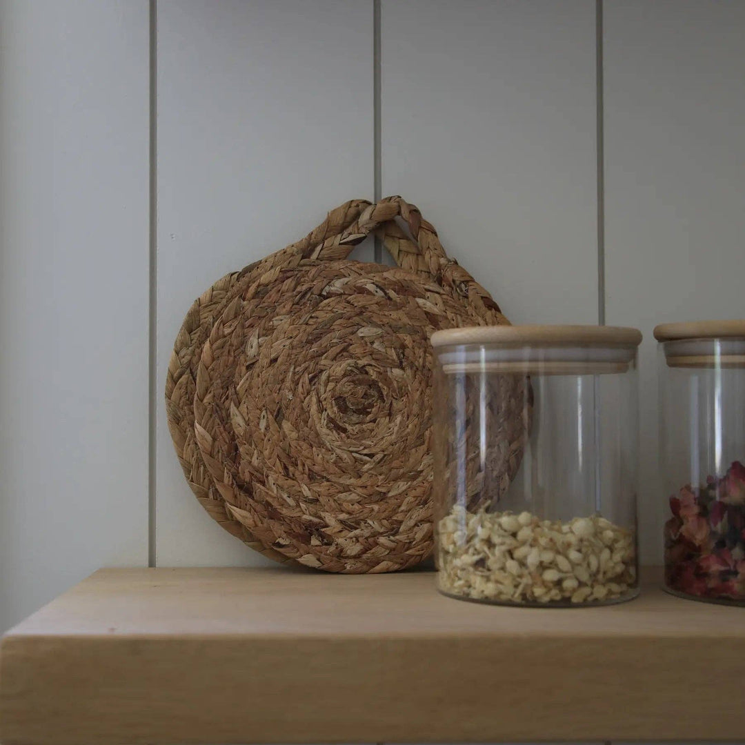 Round woven natural trivet with handle loop, placed on tabletop beside jars