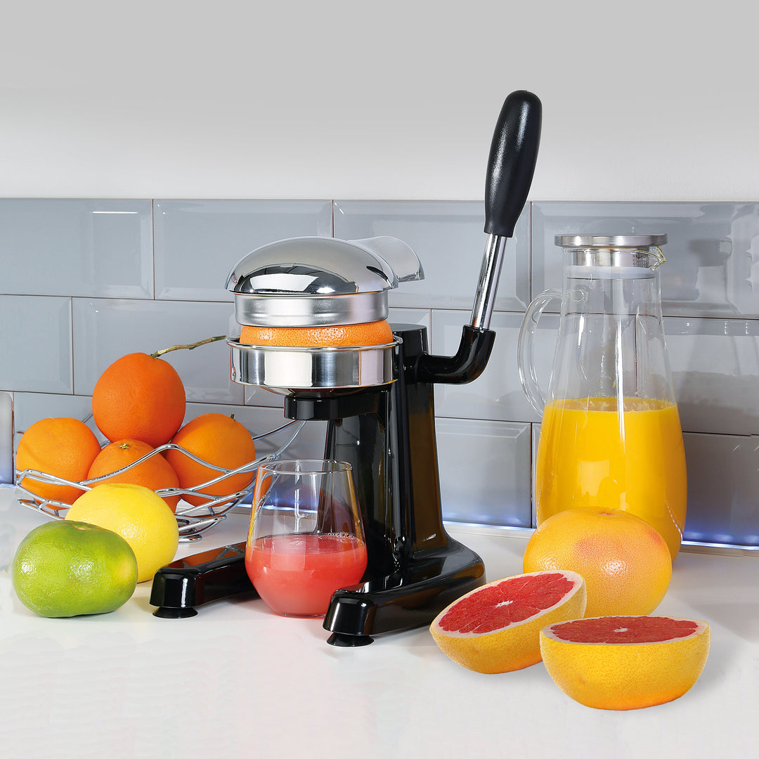 Black citrus press with chrome dome and lever, clear pitcher and cup filled with juice.