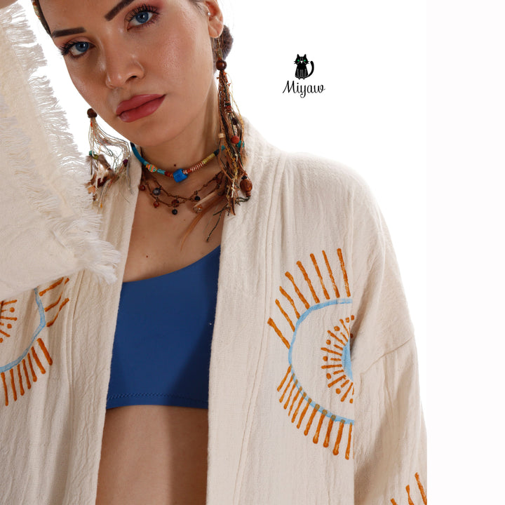 Beige cotton caftan close-up with blue evil-eye emblem, orange rays, fringe cuff