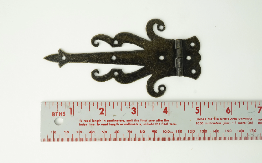 Bronze decorative strap hinge shown on a ruler scale, curled arrow-style design