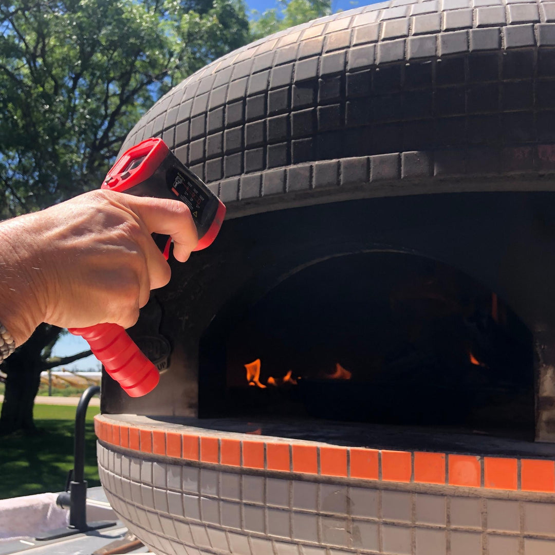 Hand holding a red-and-black infrared thermometer near a wood-fired oven opening