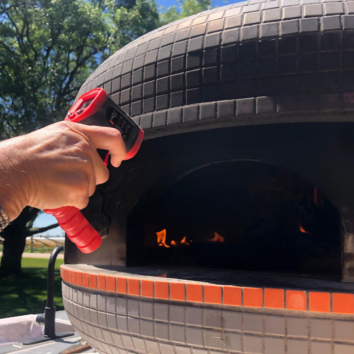 Hand holding a red-and-black infrared thermometer near a wood-fired oven opening