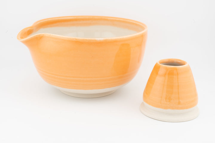 Orange ceramic matcha bowl with small spout and matching whisk stand.