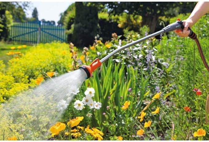 Handheld spray wand with orange trigger spraying water across a flower border.