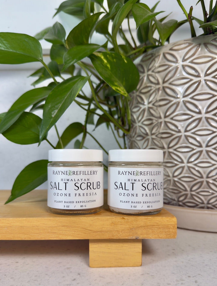 Two salt scrub jars on a wooden stand with green plant and patterned vase