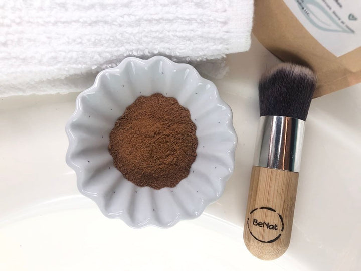 Mud mask powder in white bowl with makeup brush, beside kraft pouch