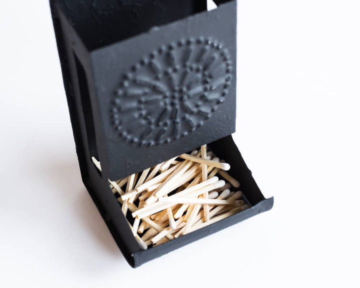 Close-up dark metal matchbox holder with embossed circular emblem and exposed matchsticks