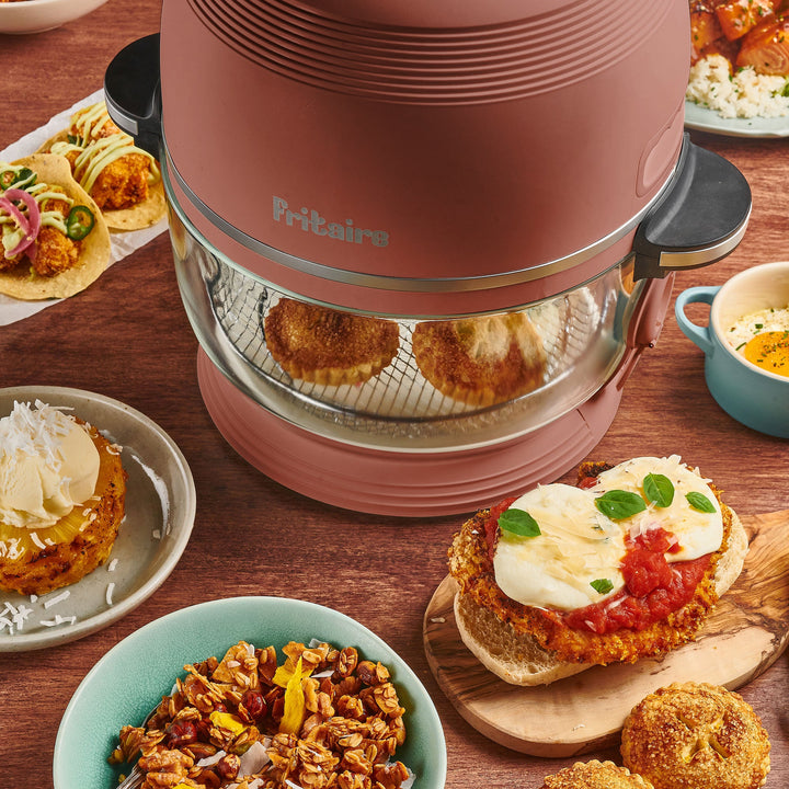 Mauve rose air fryer with glass cooking chamber beside plates of cooked dishes