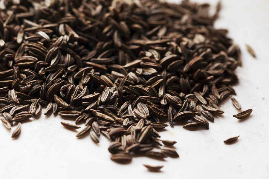 Caraway seed, whole dark brown crescent seeds scattered on a white surface.