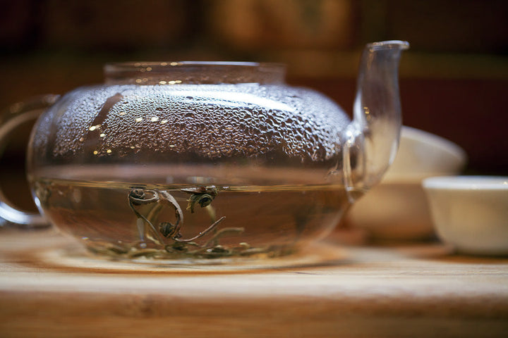 Glass teapot pouring jasmine pearl tea with visible green leaves in clear cup