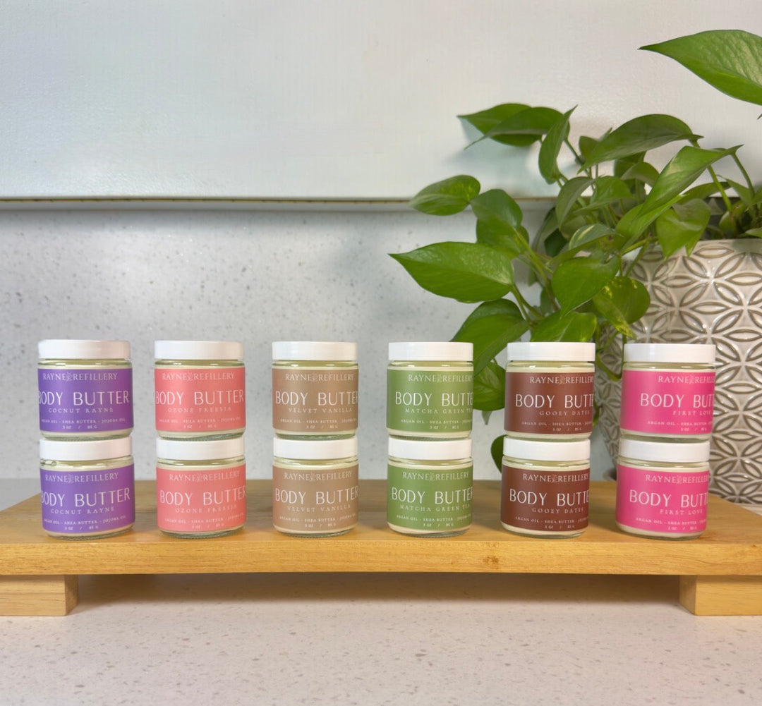 Assorted body butter jars lined up on a wooden shelf with plants