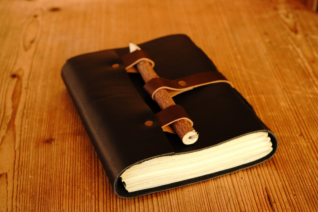 Black leather journal open with white pages and wooden pencil secured inside