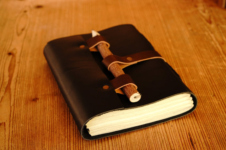 Black leather journal open with white pages and wooden pencil secured inside