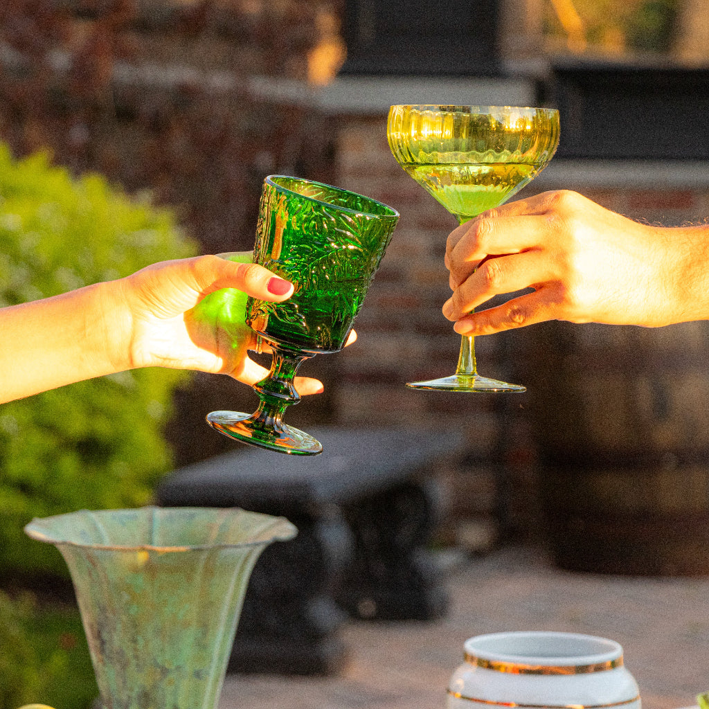 Hands clinking green botanical-imprinted goblets on outdoor patio.