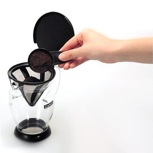 Clear glass coffee dripper with black funnel filter, showing grounds being added by hand
