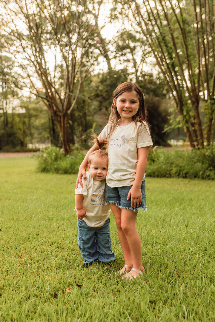 Kids organic t-shirts in cream with blue 'main squeeze' leaf graphic, worn outdoors.