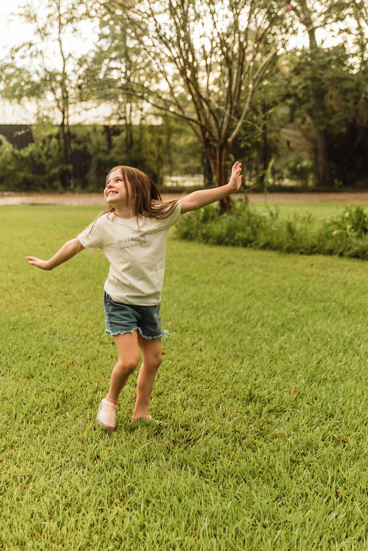 Kids organic t-shirt in cream with blue 'main squeeze' leaf graphic, worn outdoors.
