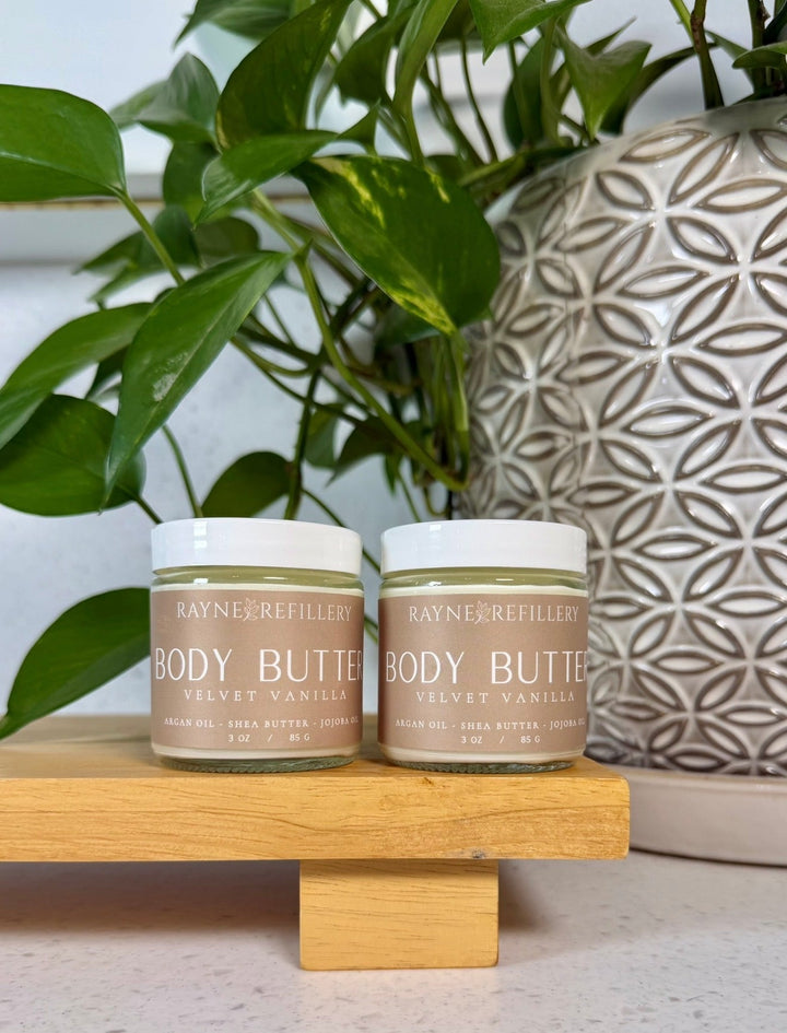 Two body butter jars with white lids on a wooden stand with greenery