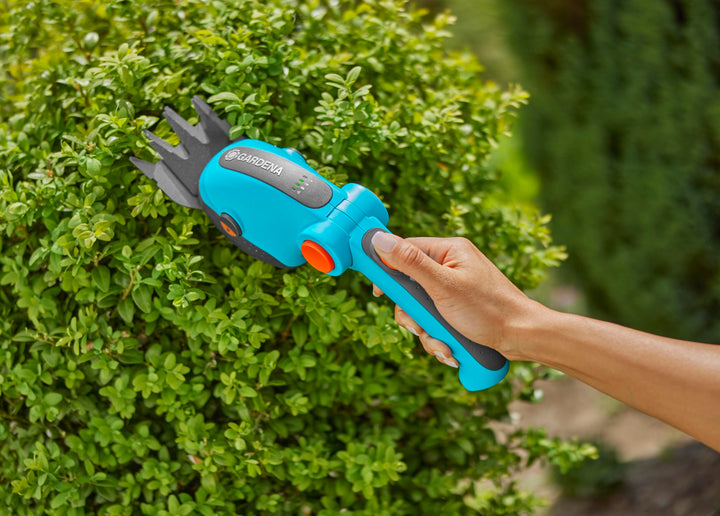Handheld teal cordless shears with orange controls cutting a green hedge