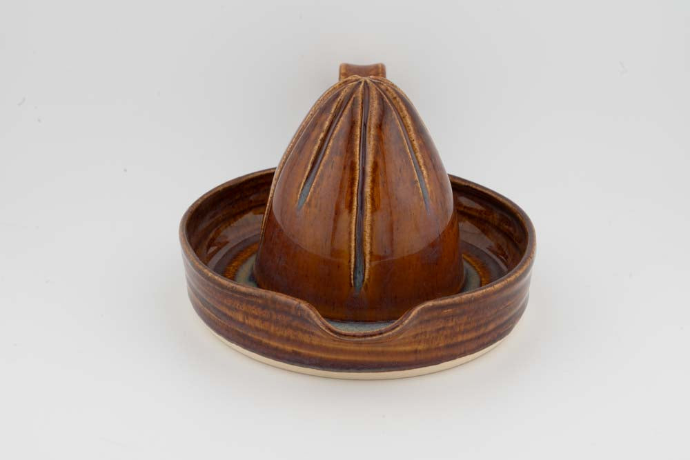 Brown ceramic citrus juicer with upright cone reamer in round drip tray; glossy glaze.