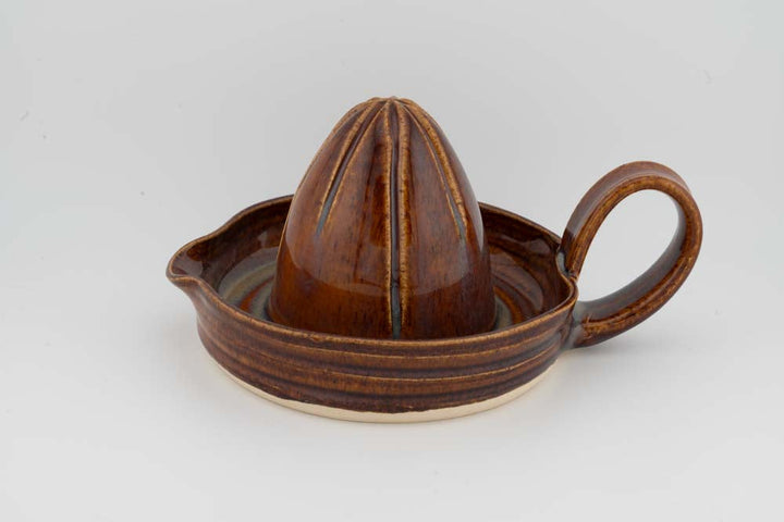 Brown ceramic citrus juicer with cone reamer, side handle, and round drip tray; glazed.