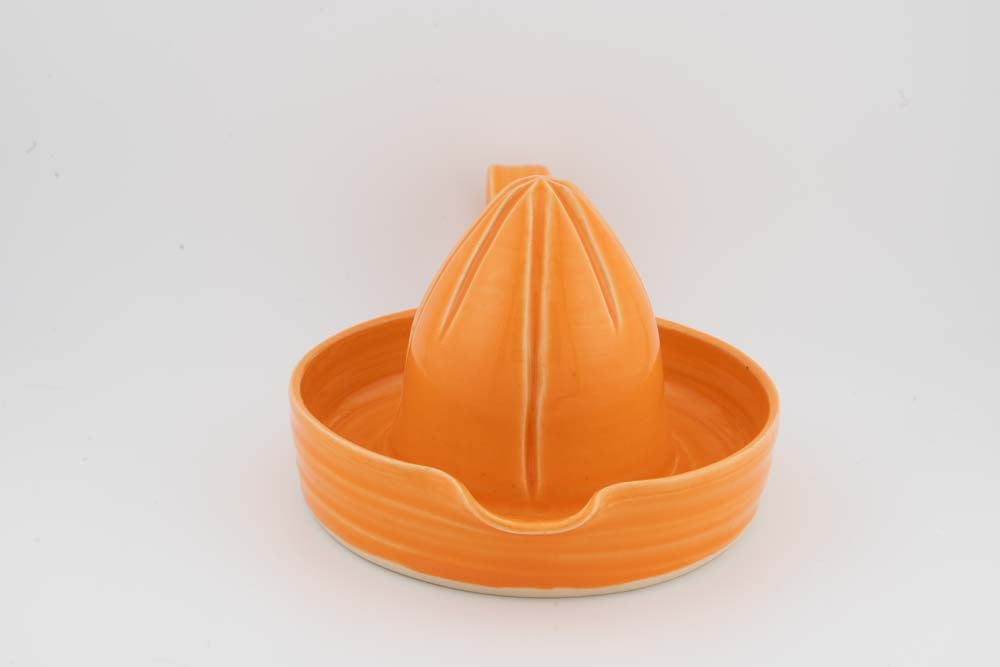 Orange ceramic citrus juicer with cone reamer in shallow drip tray; smooth glossy glaze.