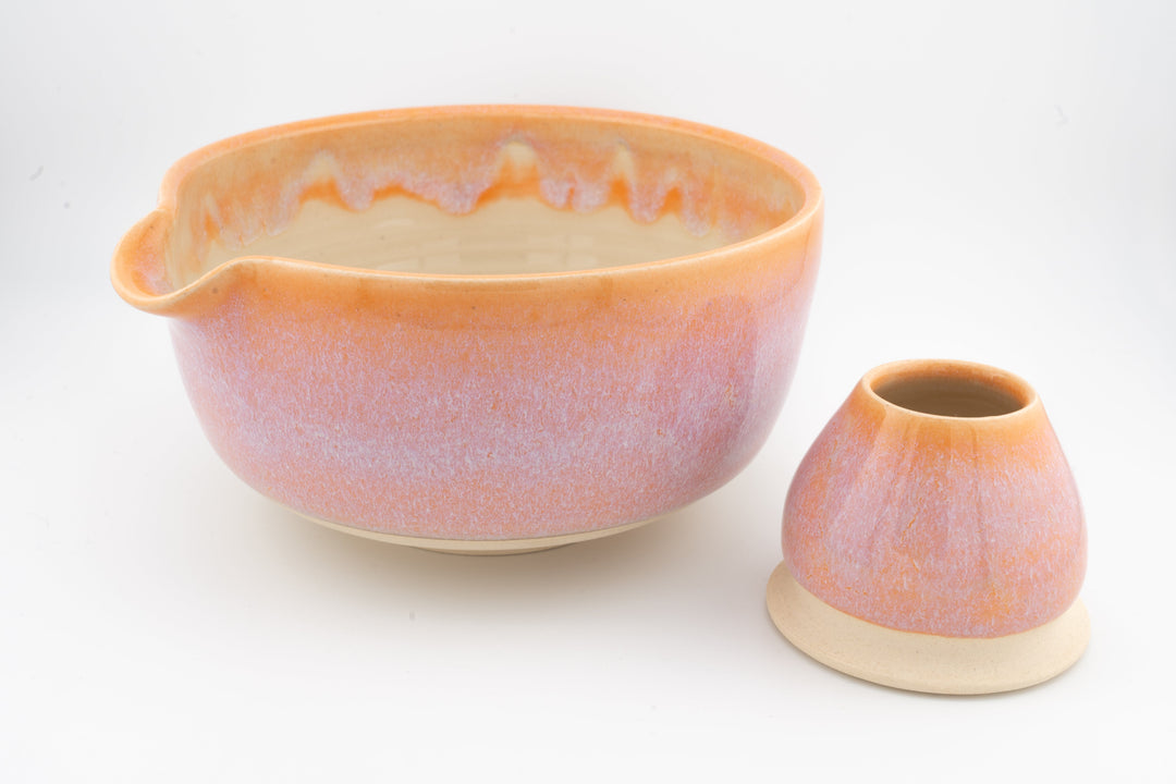 Orange-pink ceramic matcha bowl with matching whisk holder, both with speckled glaze