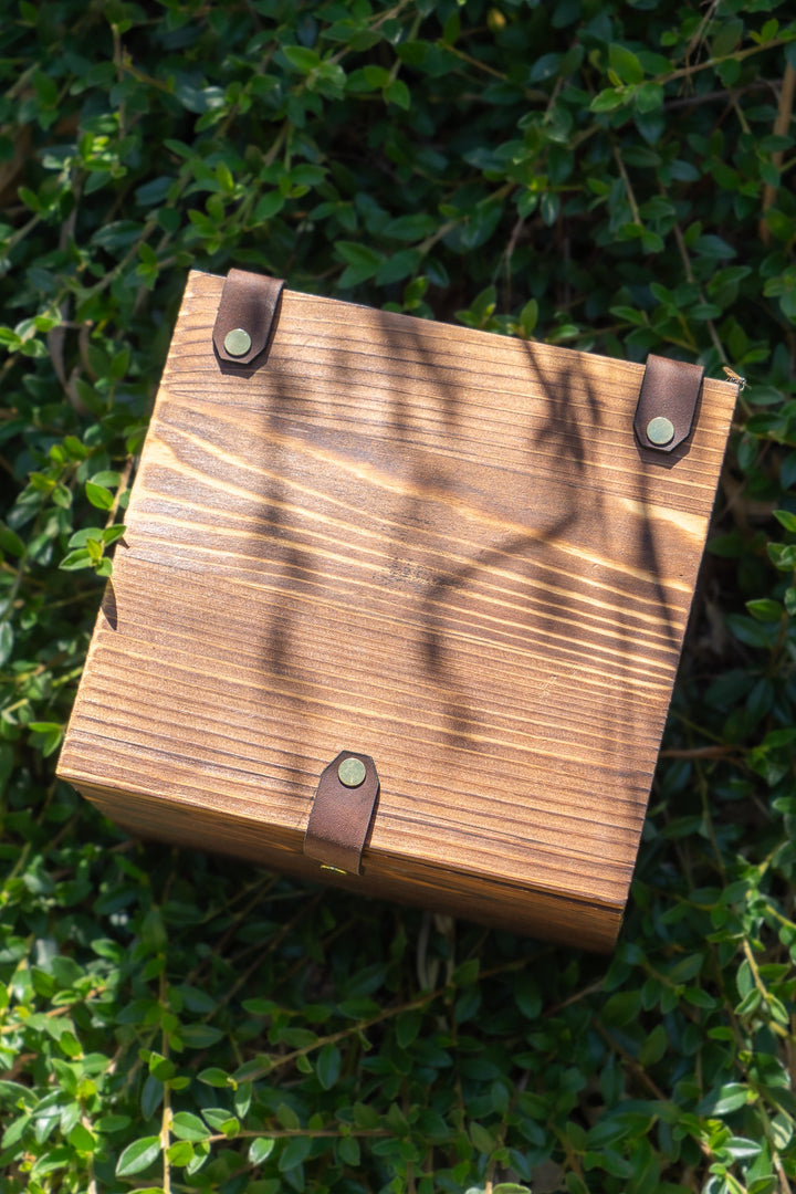 Square wooden keepsake box with metal corner studs on green outdoor foliage