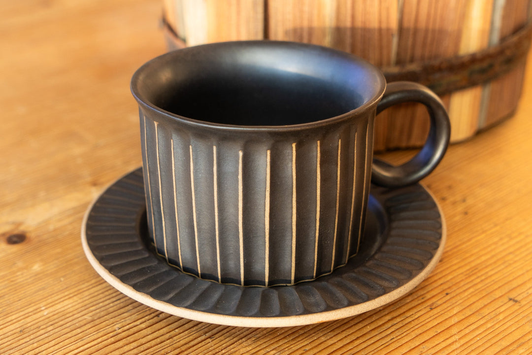 Ribbed dark mug with curved handle and matching saucer, stacked on tabletop