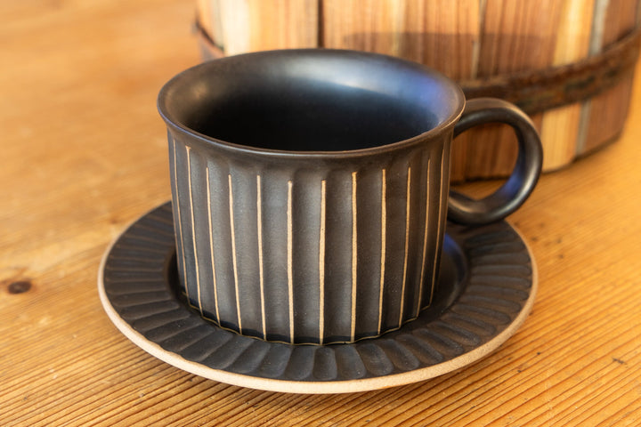 Ribbed dark mug with curved handle and matching saucer, stacked on tabletop