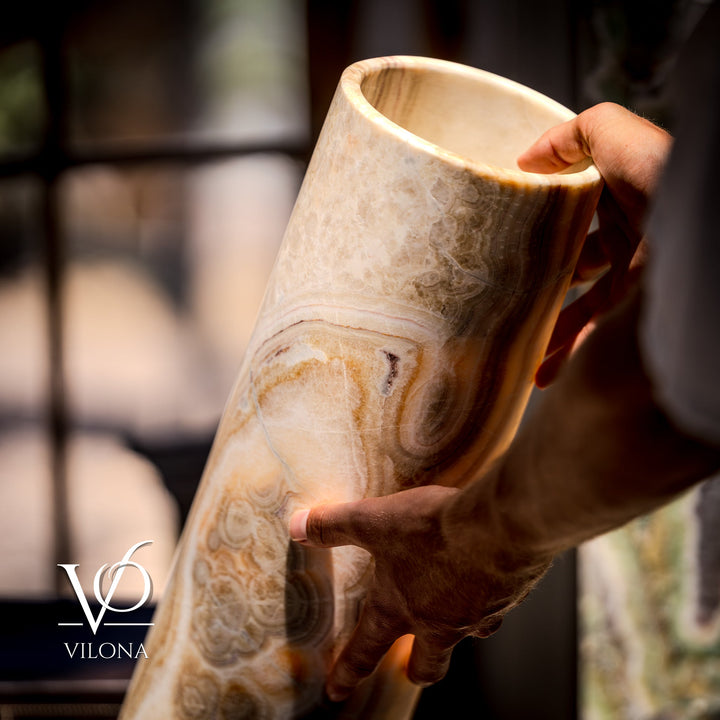 Hands holding a cream-and-brown cylindrical stone table lamp; visible natural veining.