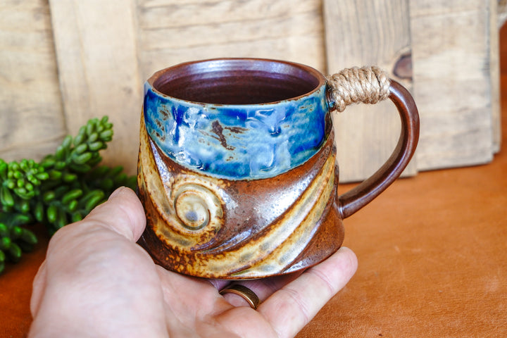 Hand holding a rustic wave ceramic mug with blue top and rope-wrapped handle