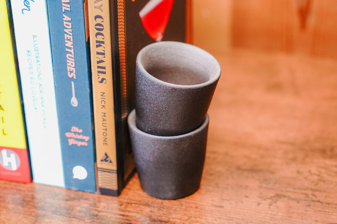 Two stacked matte stoneware shot glasses, dark speckled stoneware finish