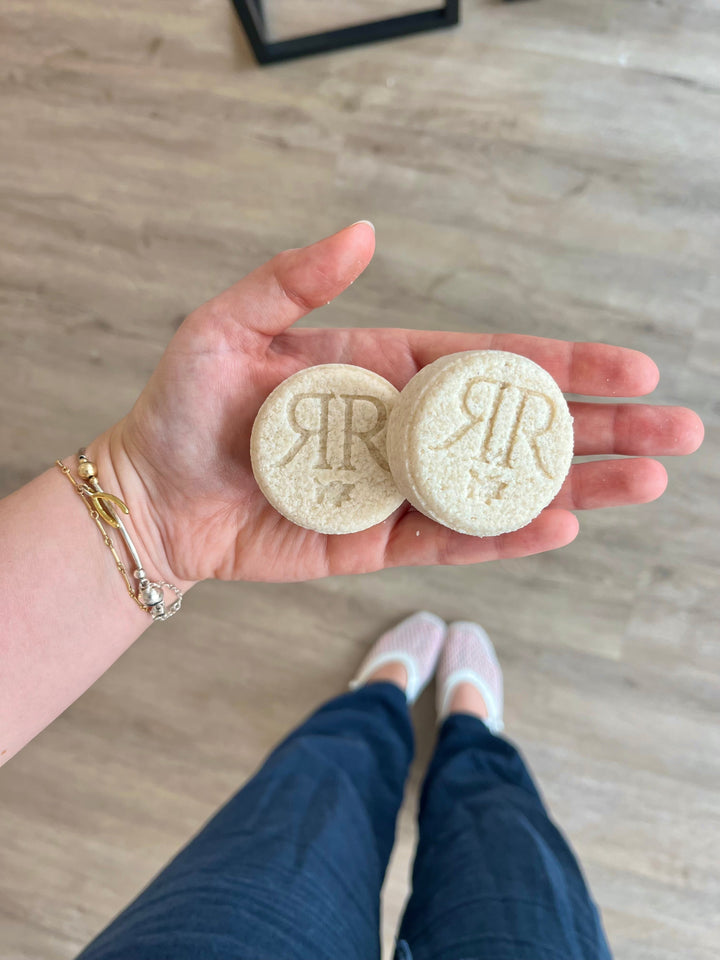 Round beige shower steamers, two tablets held in hand with embossed letters