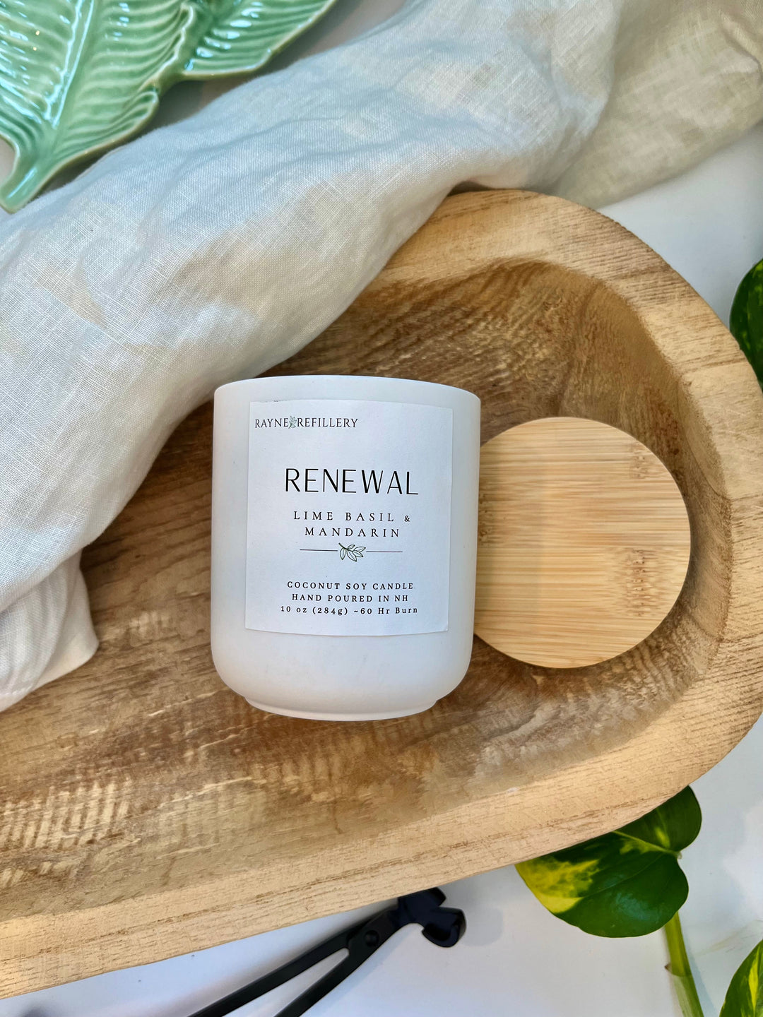 White glass coconut soy candle labeled RENEWAL with wooden lid on tray