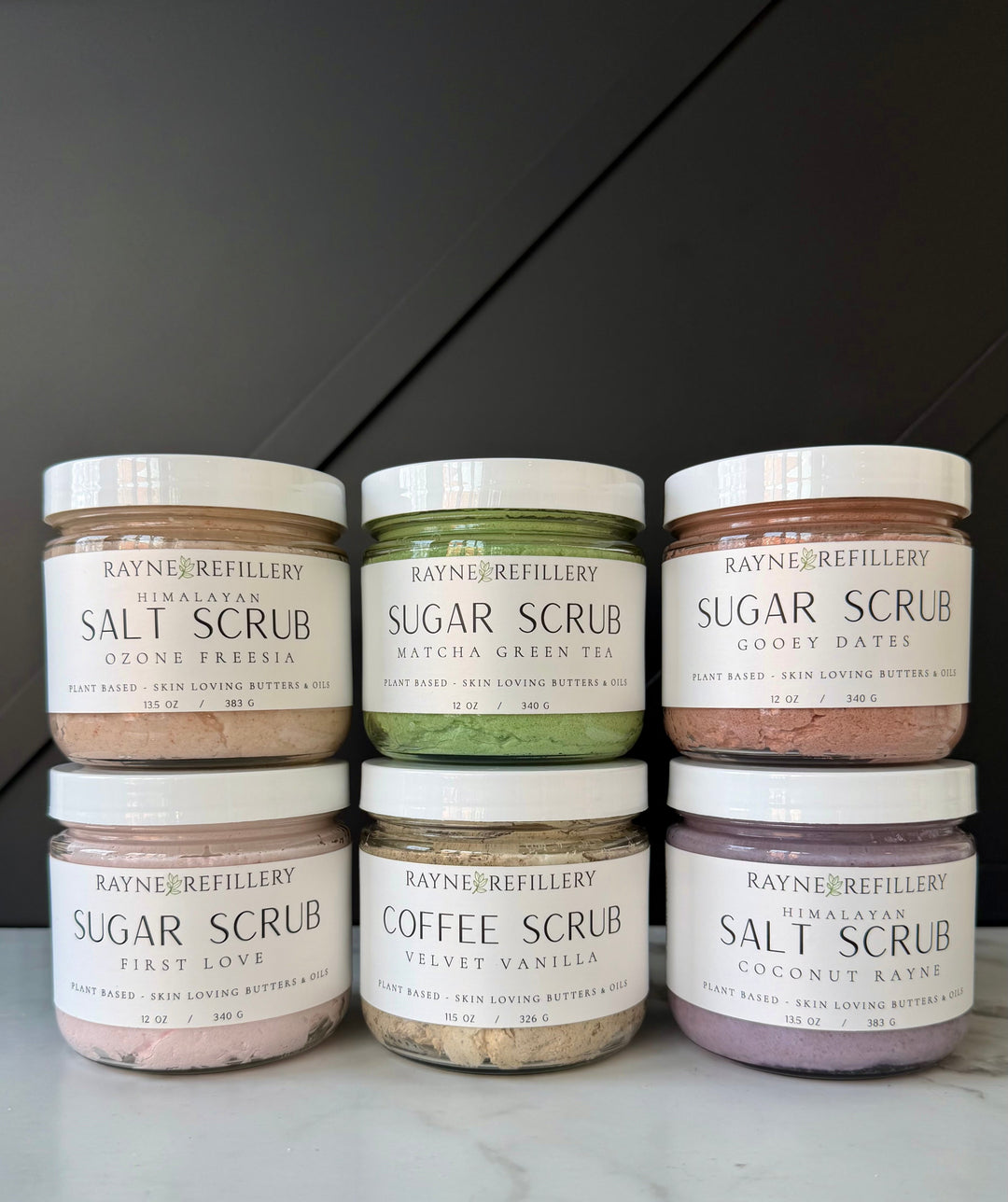 Assorted scrub jars lined up on a countertop, including salt and sugar scrubs