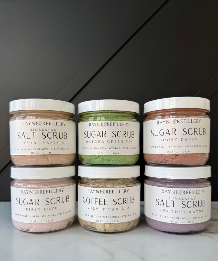 Assorted scrub jars lined up on a countertop, including salt and sugar scrubs