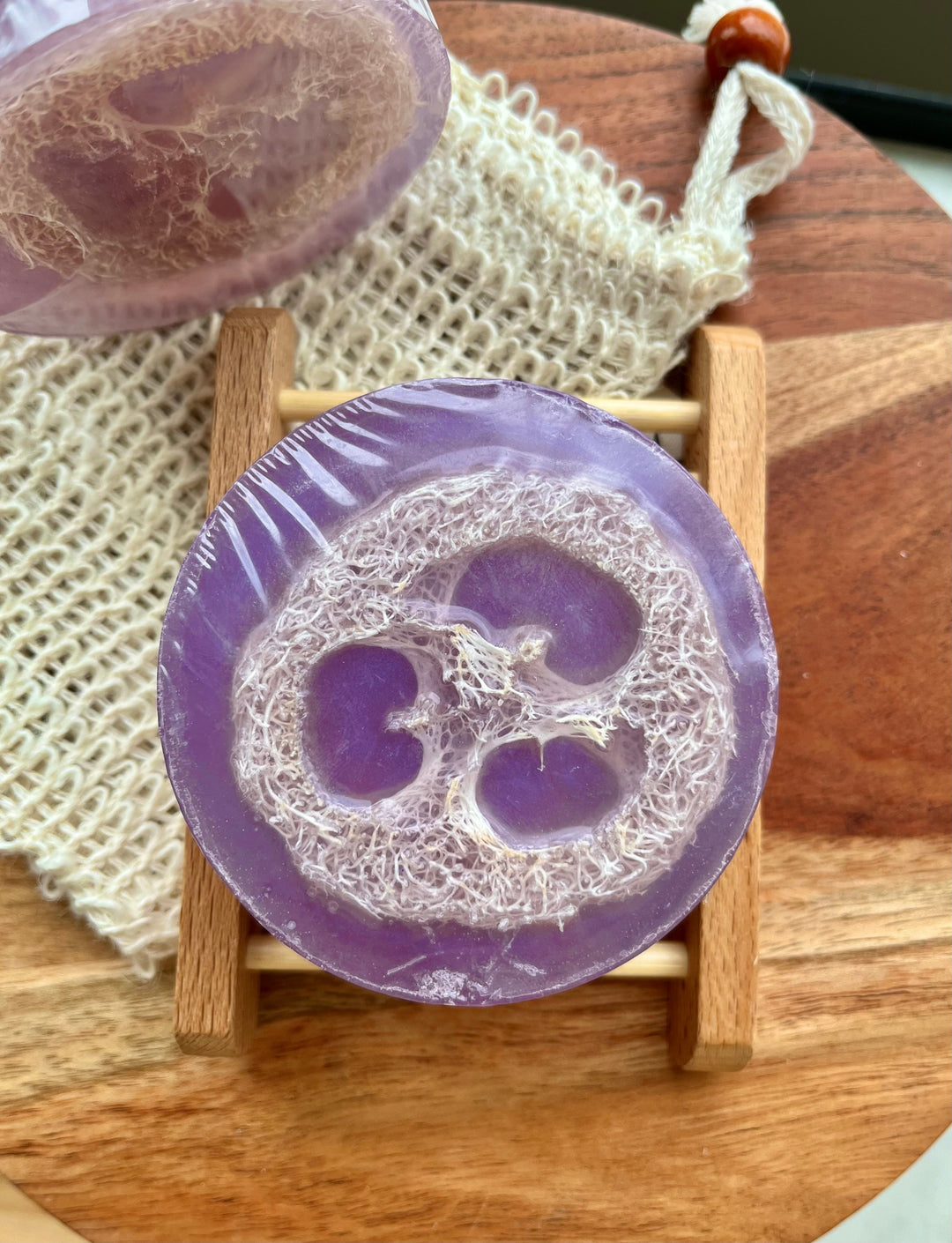 Purple loofah soap bar on small wooden stand, round with flower-shaped cutout and net bag