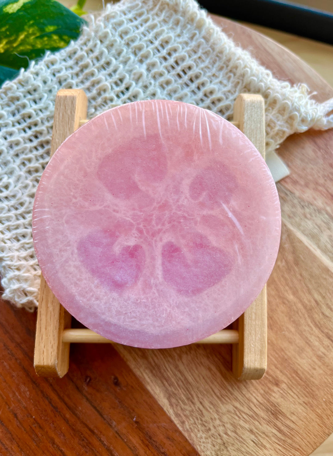 Round pink exfoliating soap bar on small wooden rack with white loofah net