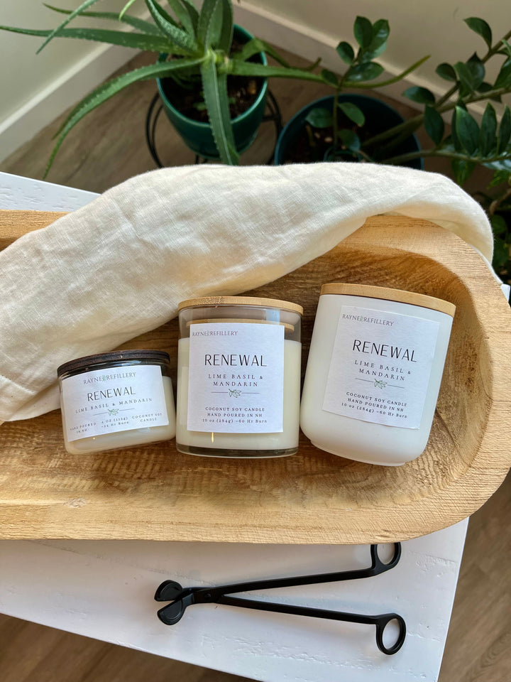 Three white glass coconut soy candles labeled RENEWAL on wooden tray