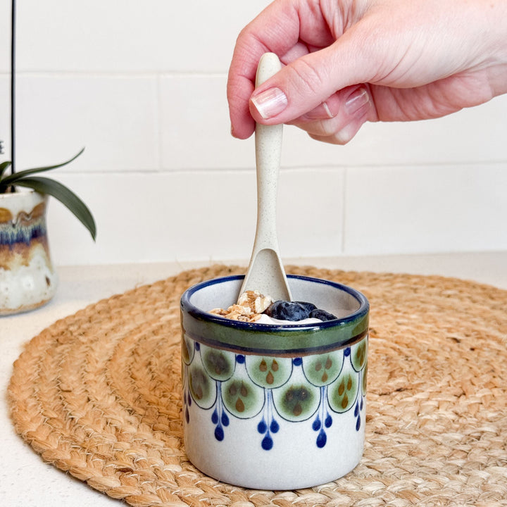 Stoneware cup on woven mat with spoon inside, green and blue raindrop pattern, blue rim.