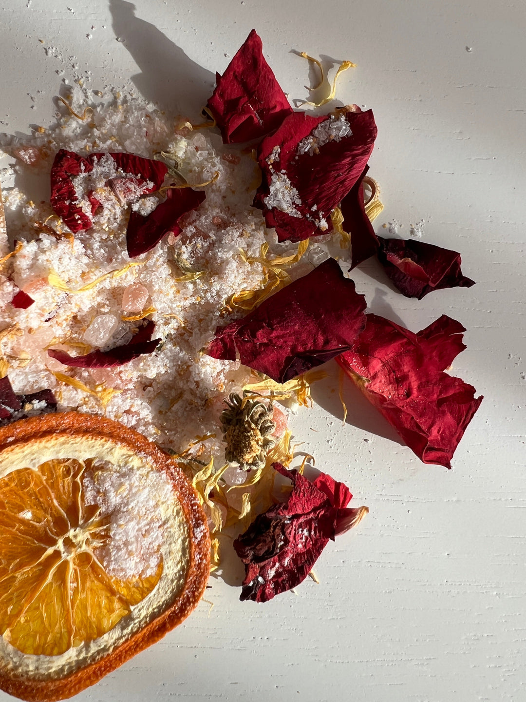 Scattered bath salts with dried rose petals and orange slice on a light surface