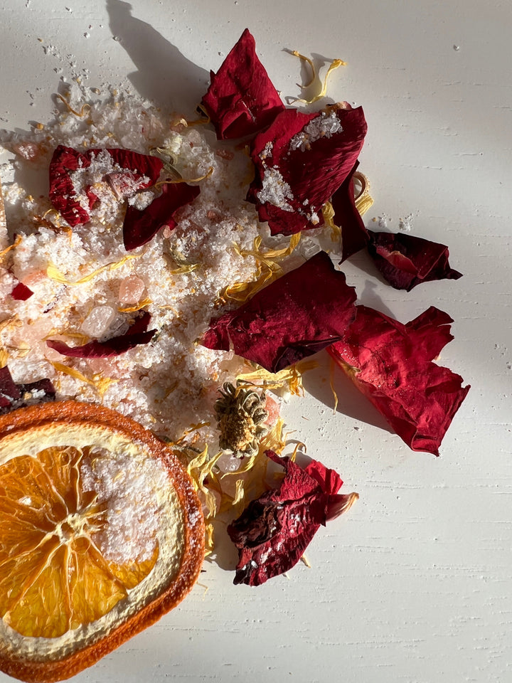 Scattered bath salts with dried rose petals and orange slice on a light surface
