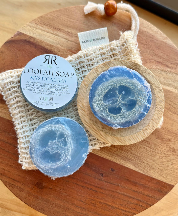 Blue loofah soap discs labeled 'Mystical Sea' on a wooden tray with a woven pouch